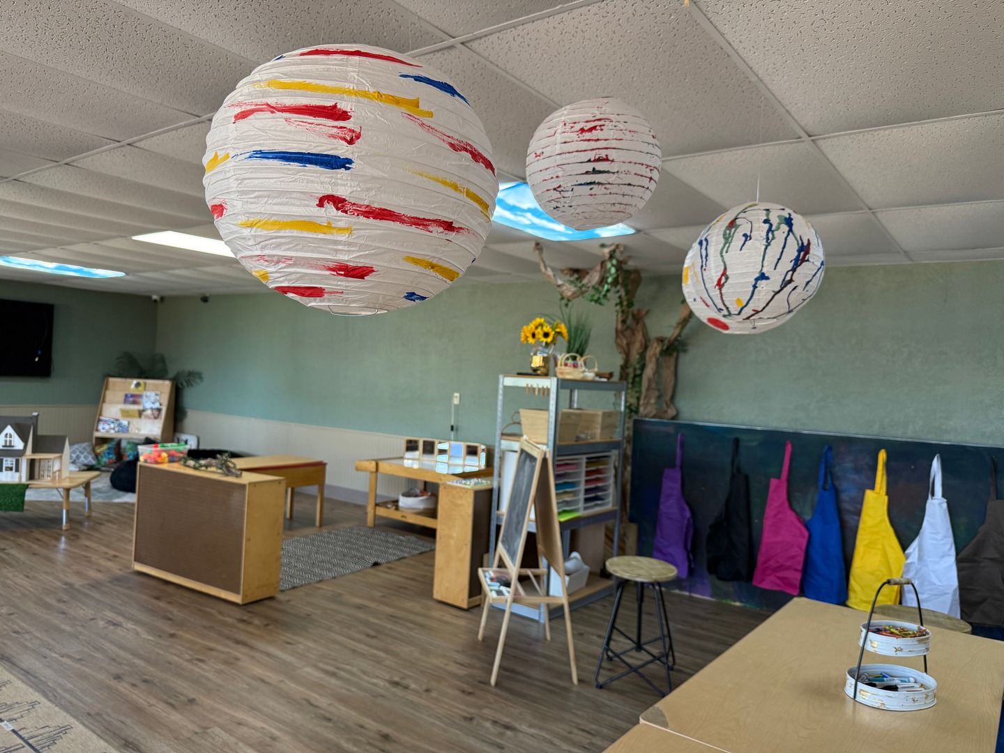 Classroom with painted paper lanterns overhead, activity tables, art aprons, and learning materials.