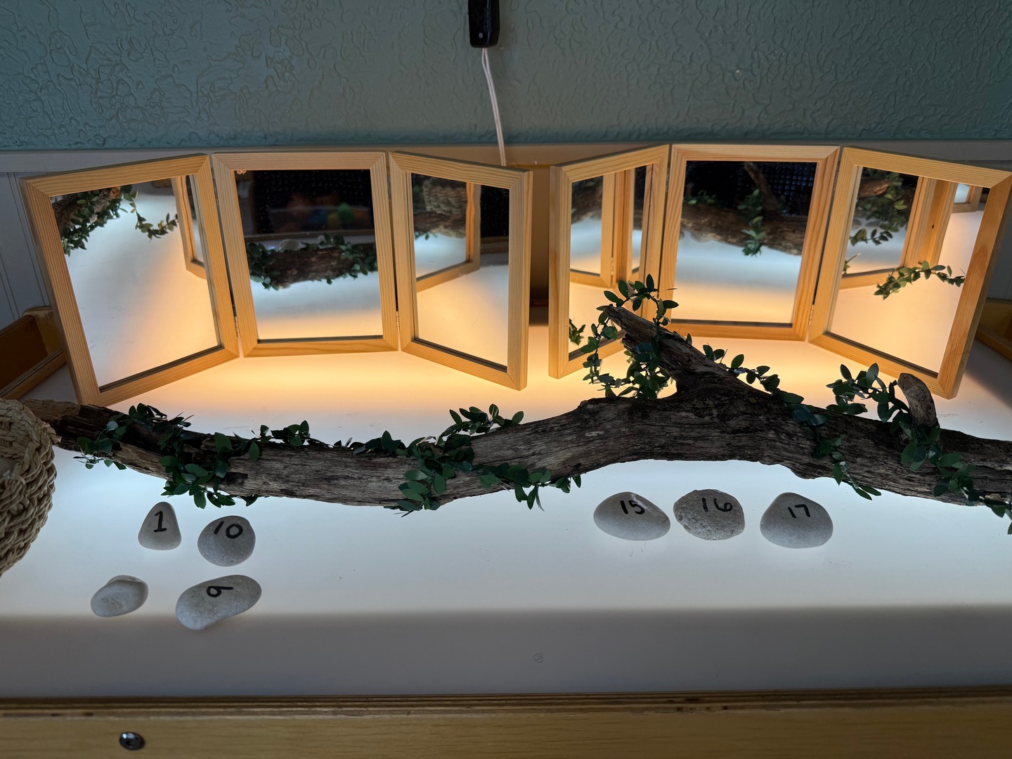 Mirrored classroom light table with a leafy branch and numbered stones arranged across the surface.