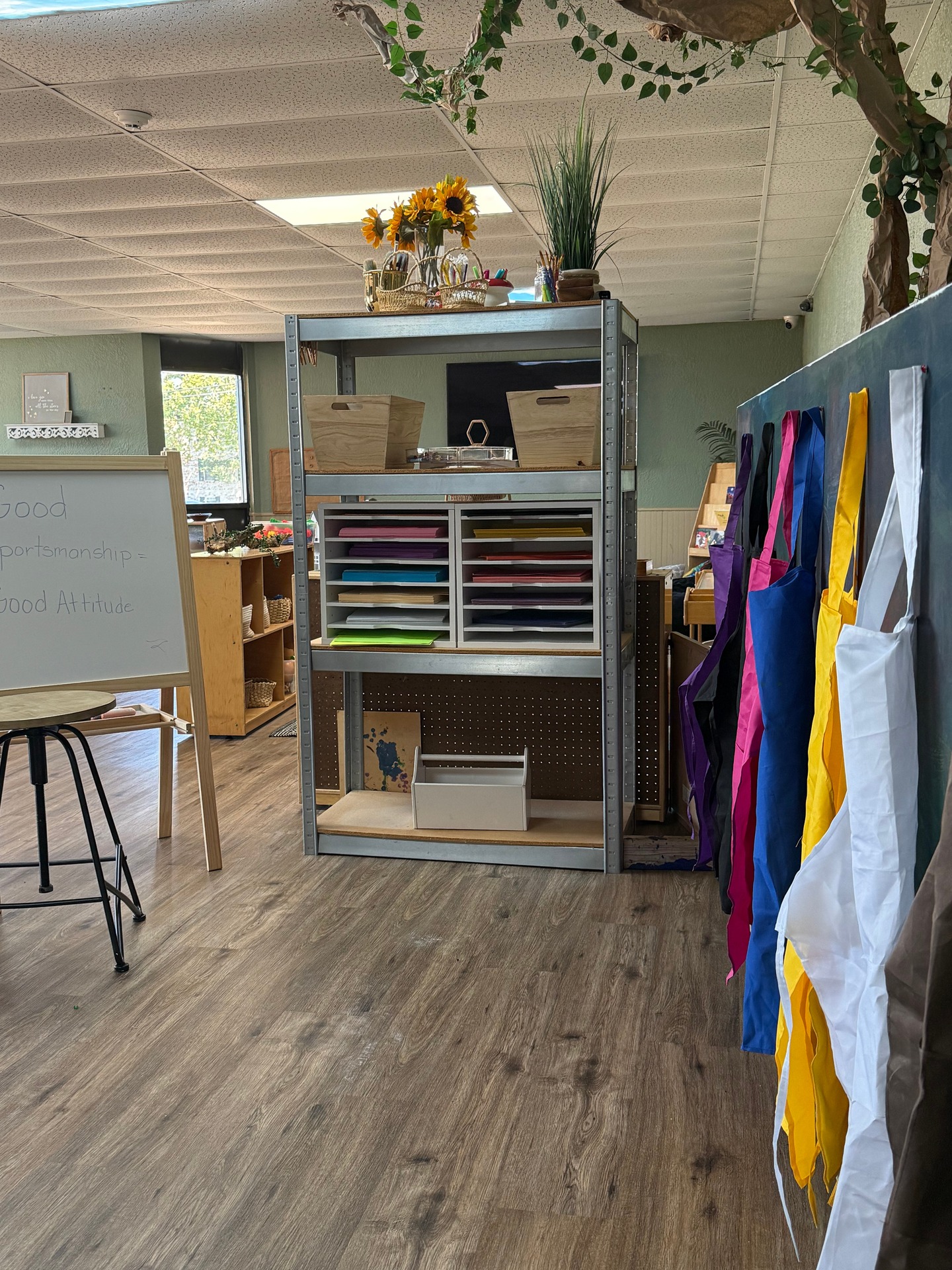 Classroom art area with organized paper trays, supply bins, sunflowers, and colorful aprons.