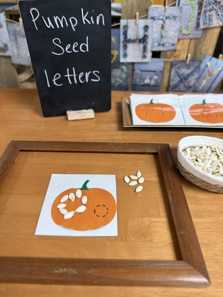 Pumpkin seeds form a letter on a pumpkin card beside a seed basket and a chalkboard labeled Pumpkin Seed Letters.