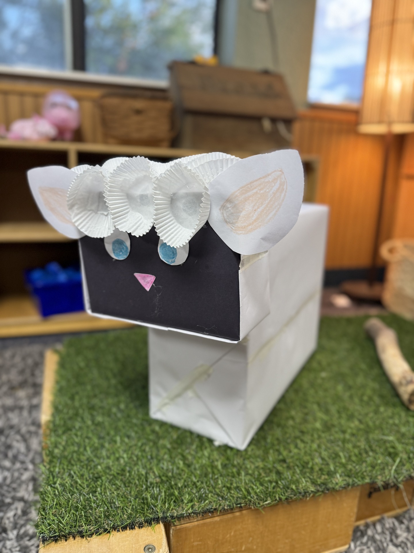 A paper-bag sheep craft with a black face, blue eyes, paper ears, and white cupcake liners on a grassy play surface.
