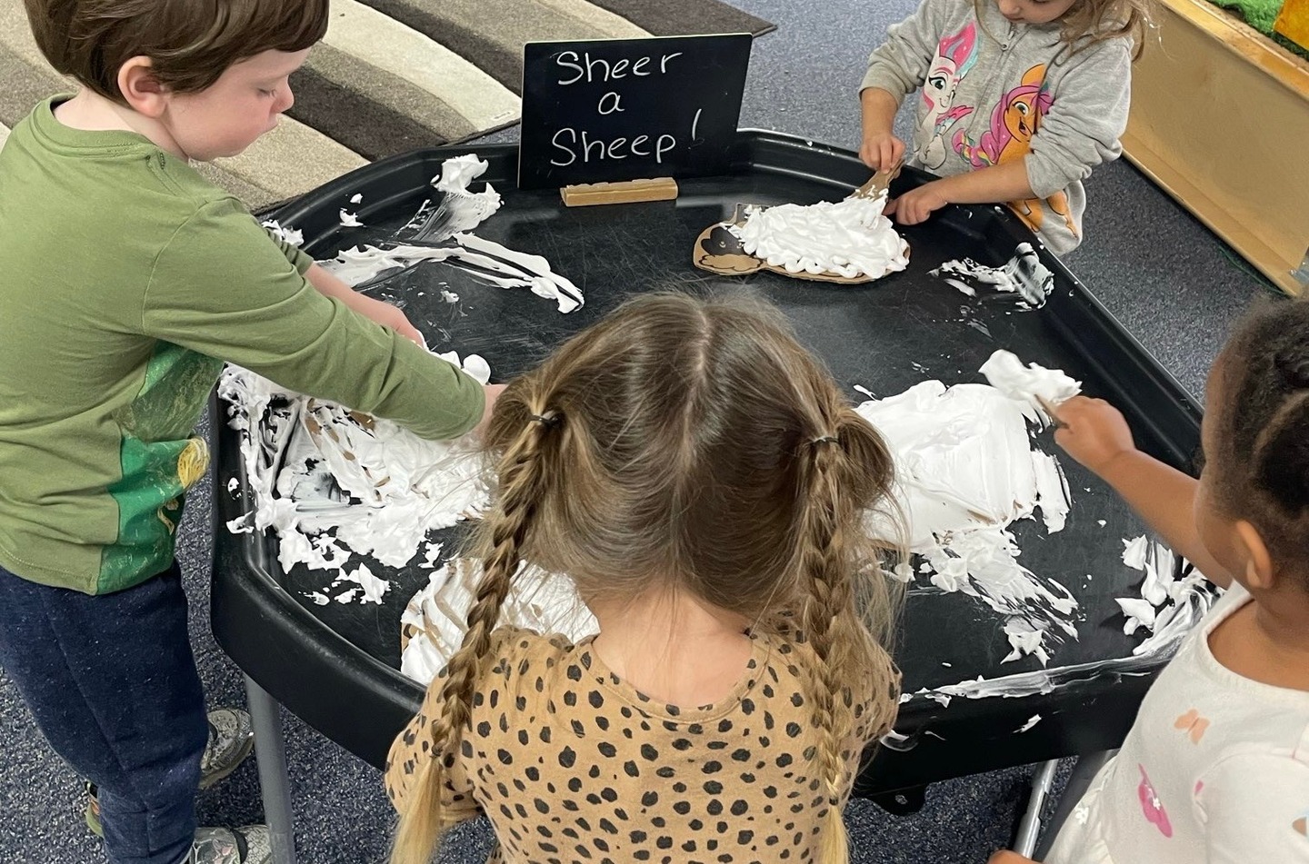 Children use wooden tools to spread white foam on sheep cutouts around a black sensory tray.