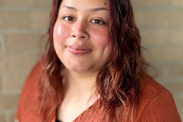 Portrait of Ms. Ivy wearing a rust-colored top against a brick wall.