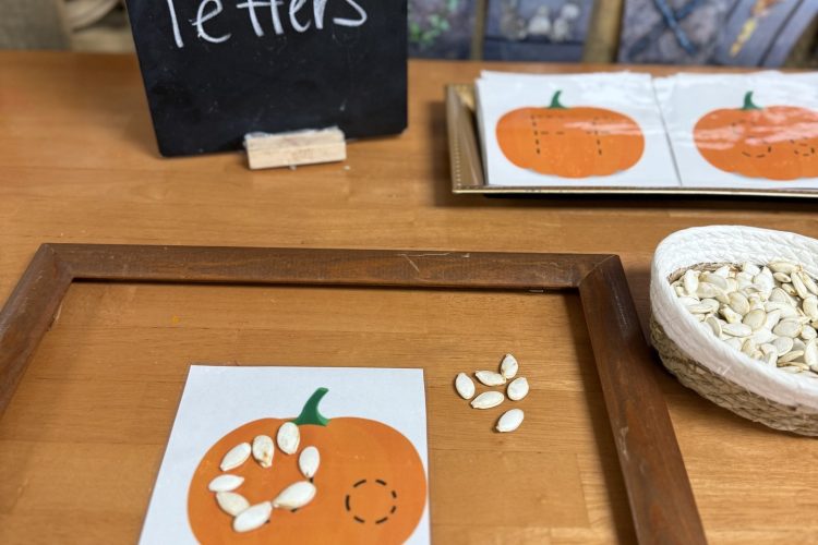 Pumpkin seeds form a letter on a pumpkin card beside a seed basket and a chalkboard labeled Pumpkin Seed Letters.